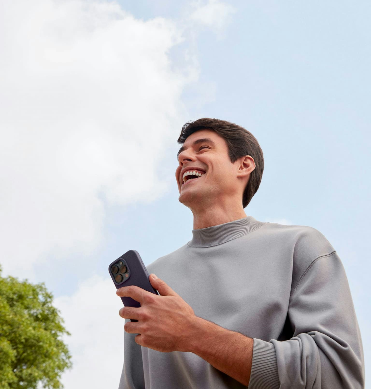 Homem branco sorrindo olhando para frente enquanto segura com a mão esquerda um celular, em um ambiente externo a luz do dia com nuvens no céu e uma árvore a sua esquerda e outra árvore a sua direita.