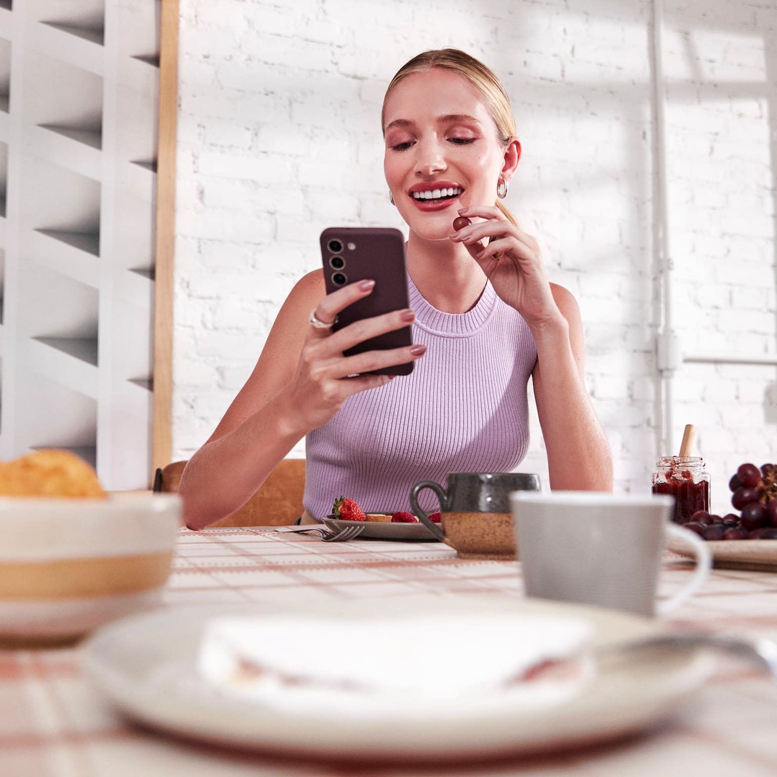 Mulher branca com o cabelo preso, sentada e sorrindo enquanto olha para o celular em uma de suas mãos e com a outra leva uma uva a sua boca, a sua frente uma mesa com alguns pratos, xícaras, tigelas com frutas e uma jarra de suco ao lado. Ao fundo uma parede de tijolos brancos com um quadro abstrato iluminados pela luz do dia de parte e uma janela.