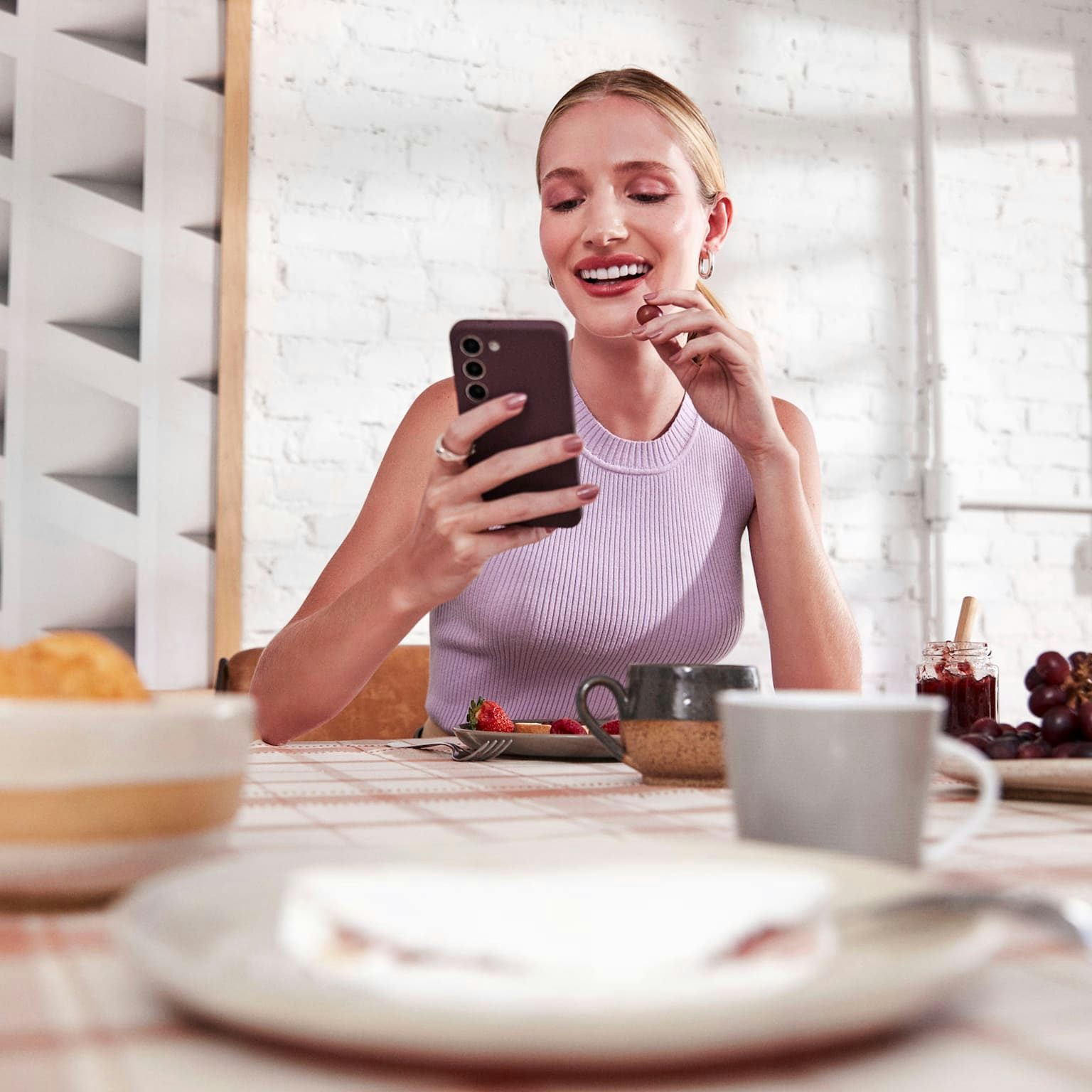 Mulher branca com o cabelo preso, sentada e sorrindo enquanto olha para o celular em uma de suas mãos e com a outra leva uma uva a sua boca, a sua frente uma mesa com alguns pratos, xícaras, tigelas com frutas e uma jarra de suco ao lado. Ao fundo uma parede de tijolos brancos com um quadro abstrato iluminados pela luz do dia de parte e uma janela.