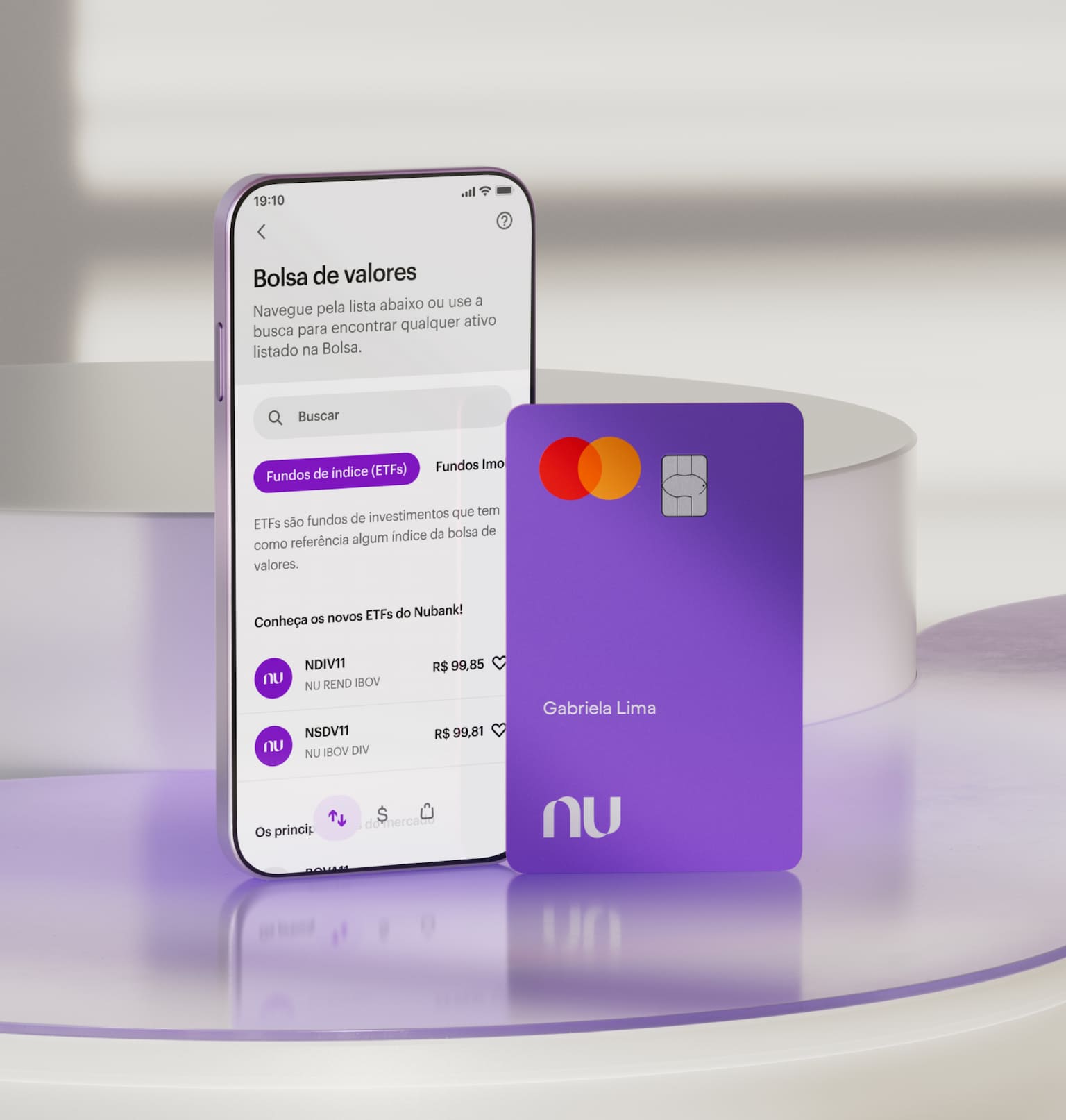 Celular com aplicativo Nubank aberto na área de Investimentos em Bolsa de Valores com opções para investir na parte superior da tela. Na parte inferior do aplicativo na tela do celular, sugestões de novos ETFs do Nubank. O celular e o cartão estão em pé apoiados em uma forma geométrica branca.