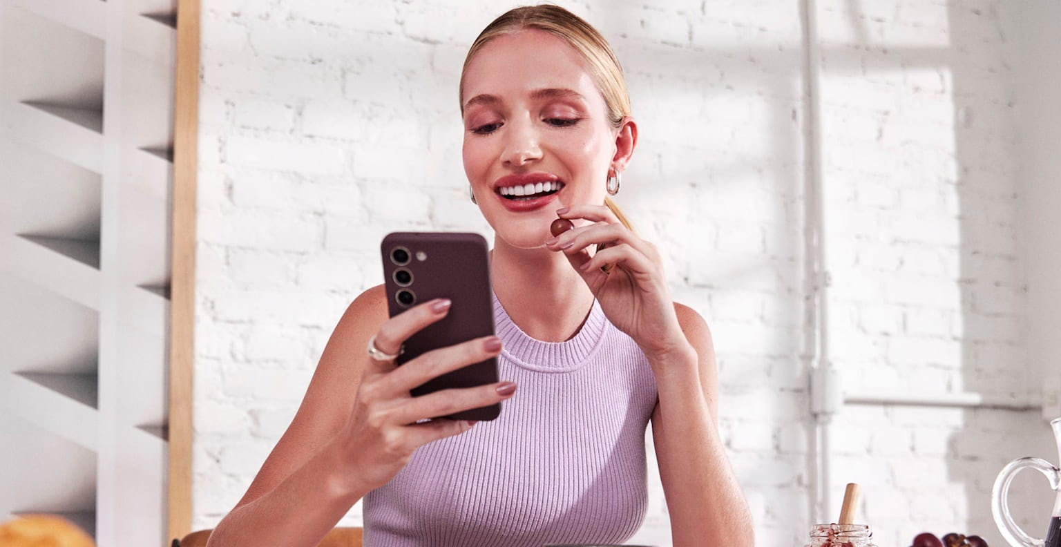 Mulher branca com o cabelo preso, sentada e sorrindo enquanto olha para o celular em uma de suas mãos e com a outra leva uma uva a sua boca, a sua frente uma mesa com alguns pratos, xícaras, tigelas com frutas e uma jarra de suco ao lado. Ao fundo uma parede de tijolos brancos com um quadro abstrato iluminados pela luz do dia de parte e uma janela.