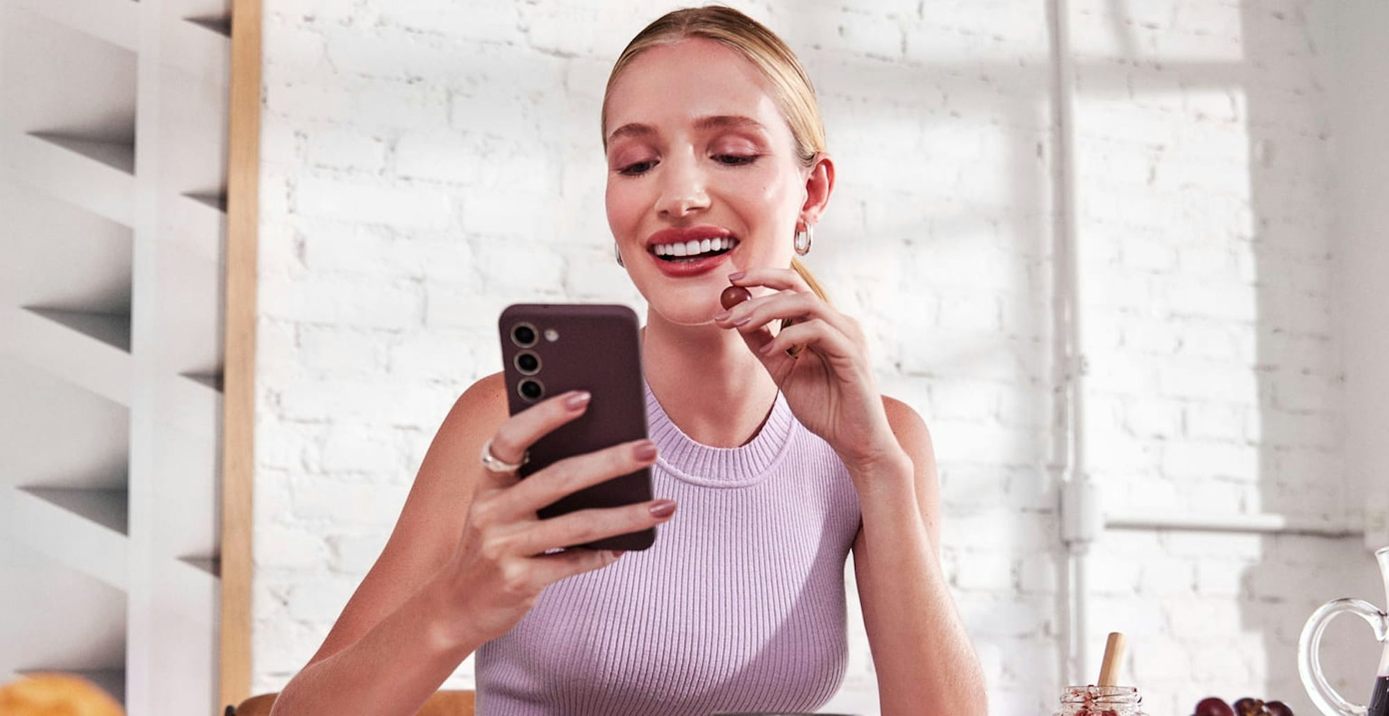 Mulher branca com o cabelo preso, sentada e sorrindo enquanto olha para o celular em uma de suas mãos e com a outra leva uma uva a sua boca, a sua frente uma mesa com alguns pratos, xícaras, tigelas com frutas e uma jarra de suco ao lado. Ao fundo uma parede de tijolos brancos com um quadro abstrato iluminados pela luz do dia de parte e uma janela.