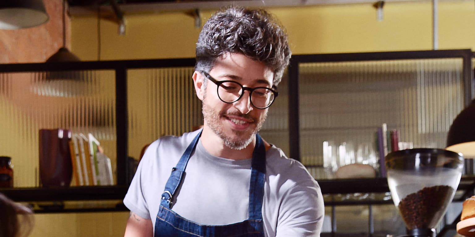 Homem branco de óculos vestido com uma camiseta cinza e por cima um avental azul olhando para baixo estende o braço esquerdo.