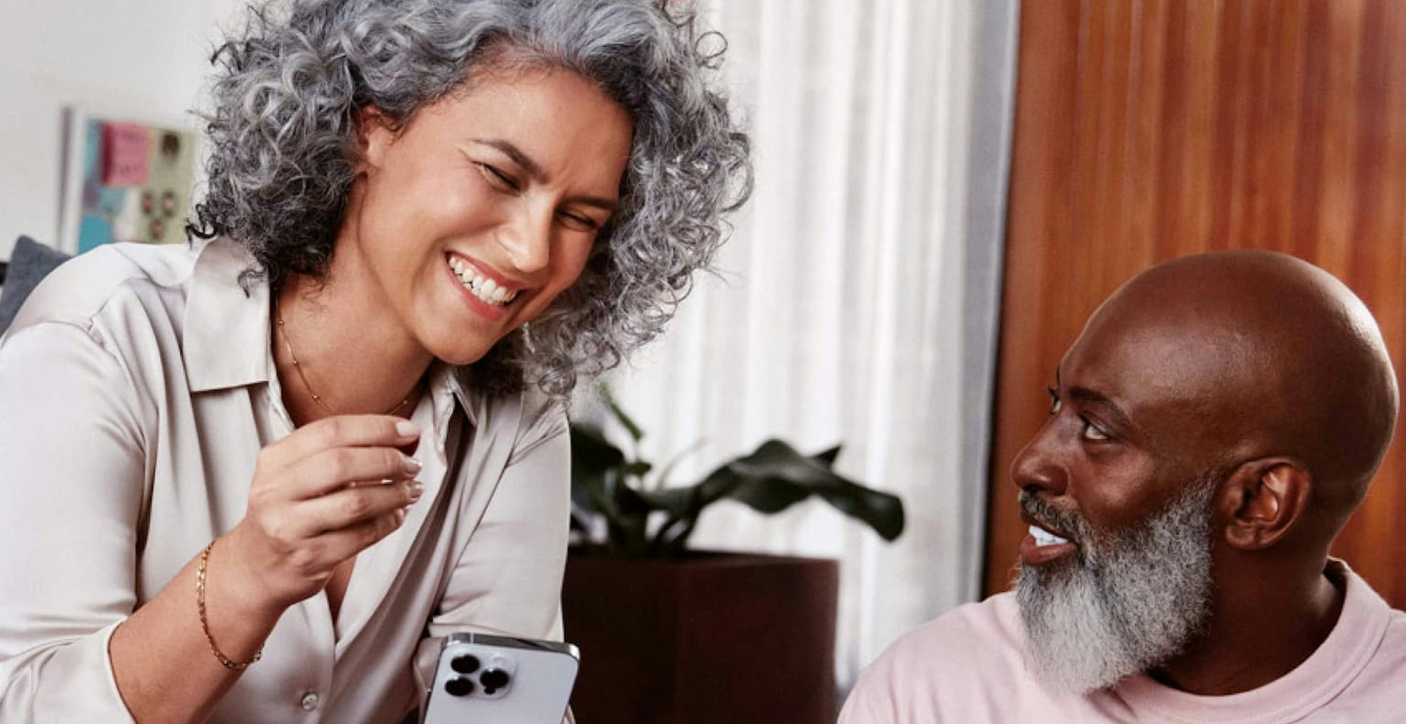 Mulher negra de cabelos cacheados e grisalhos sentada em um sofá e sorrindo, segurando com a mão esquerda um celular em direção a um homem negro, careca, com barba grisalha, sentado ao seu lado no chão, olhando para o rosto dela enquanto sorri e com uma pequena mesa de madeira a sua frente com um notebook.