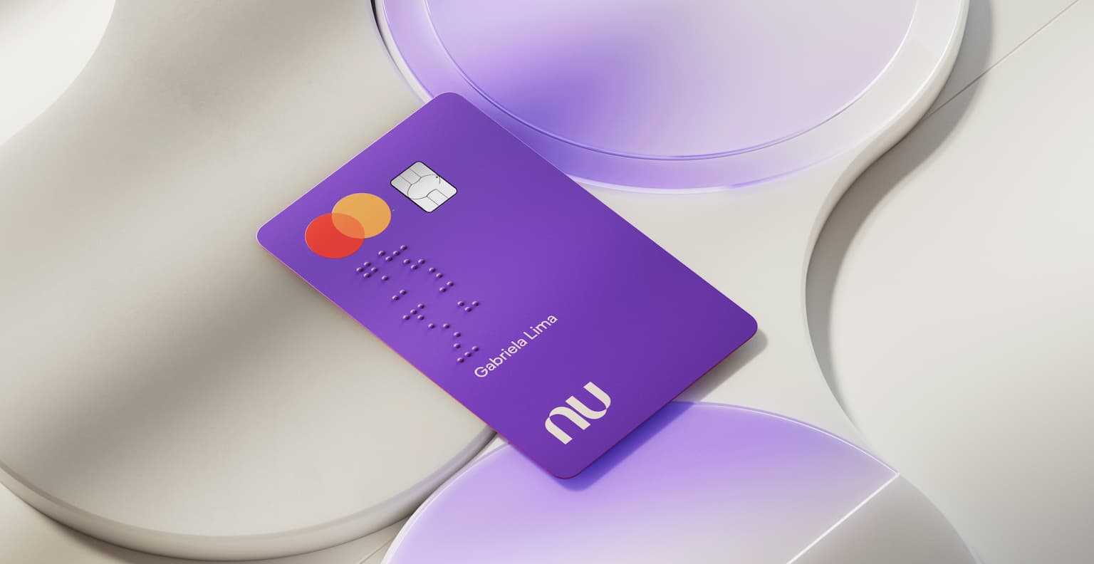 Cartão do Nubank visto de cima posto sob superfícies geométricas circulares. O cartão está com a face principal virada para cima, com o logo da bandeira Mastercard, chip, nome do proprietário do cartão escrito e também em braille, acima do logo do Nubank.