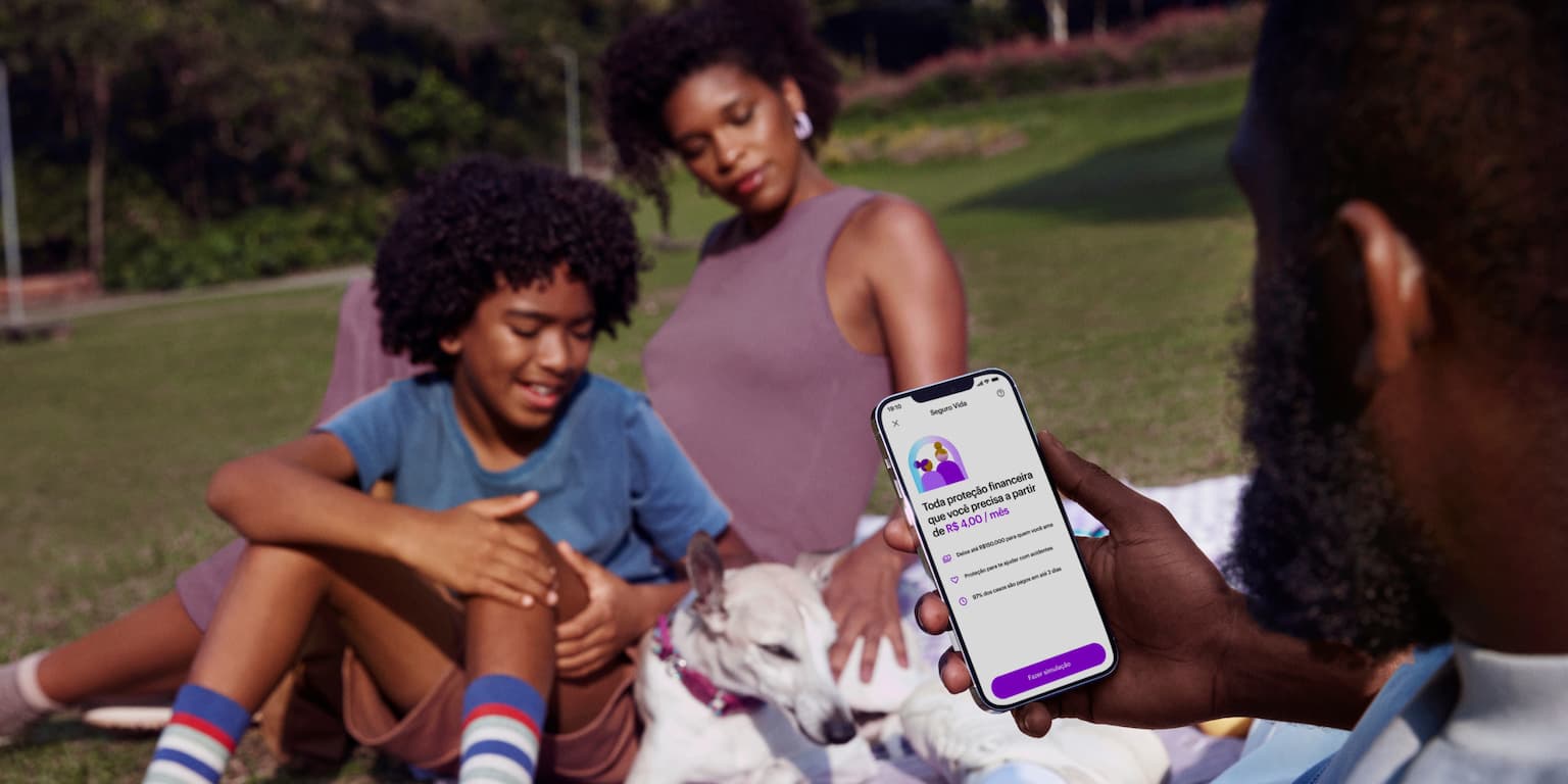 Homem sentado segurando um celular com o aplicativo do Nubank aberto. Ao fundo está sua família e seu cachorro. Todos estão sentados no chão, em cima de um pano com detalhes roxos, onde também há uma câmera fotográfica roxa.