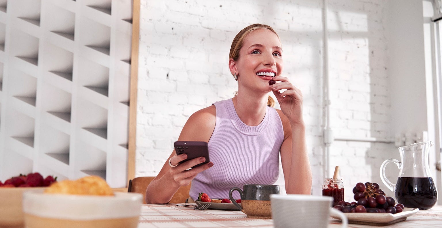 Mulher branca com o cabelo preso, sentada, sorrindo e olhando para frente enquanto segura o celular em uma de suas mãos e com a outra leva uma uva a sua boca, a sua frente uma mesa com alguns pratos, xícaras, tigelas com frutas e uma jarra de suco ao lado. Ao fundo uma parede de tijolos brancos com um quadro abstrato iluminados pela luz do dia e parte de uma janela.