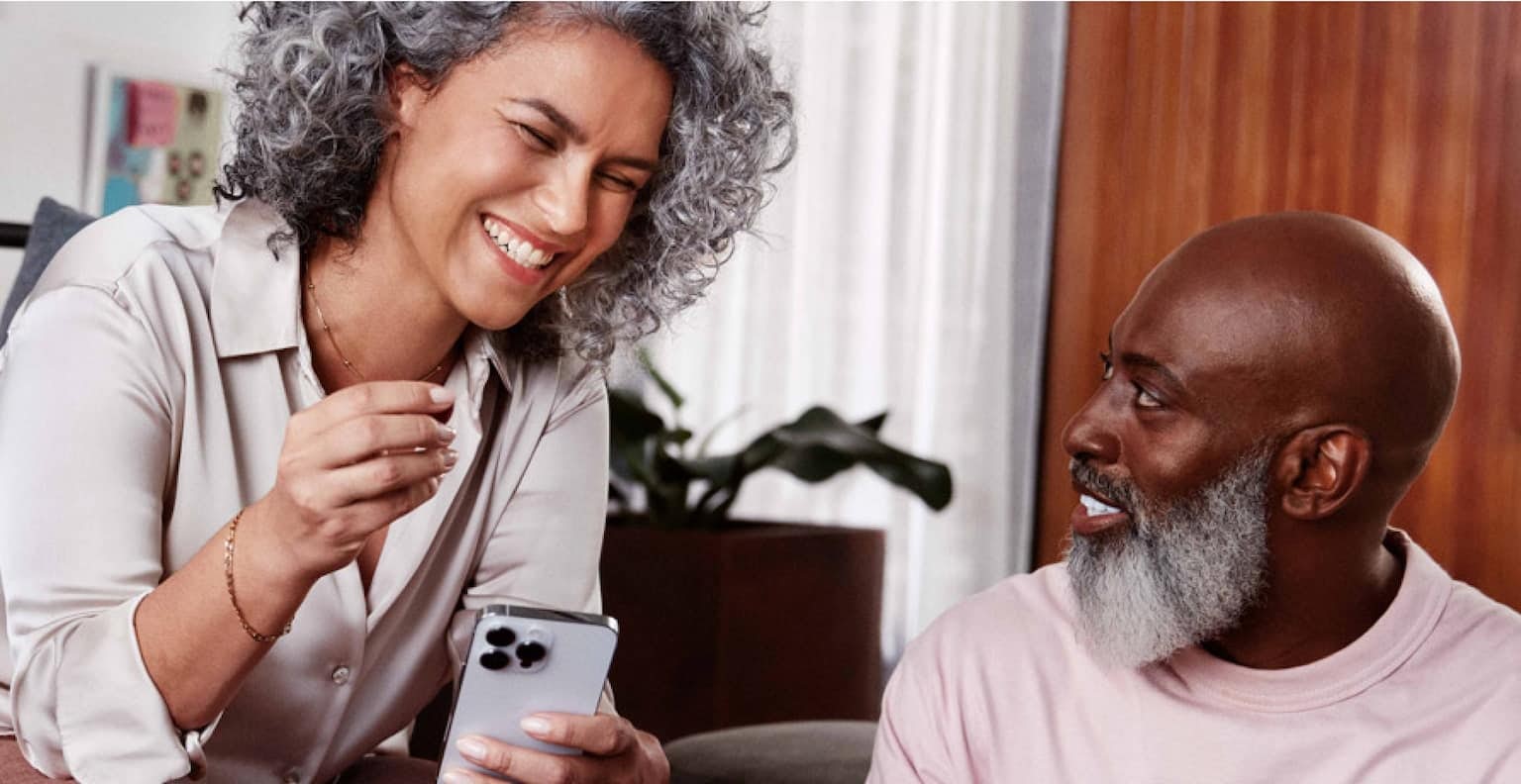 Mulher negra de cabelos cacheados e grisalhos sentada em um sofá e sorrindo, segurando com a mão esquerda um celular em direção a um homem negro, careca, com barba grisalha, sentado ao seu lado no chão, olhando para o rosto dela enquanto sorri e com uma pequena mesa de madeira a sua frente com um notebook. Em destaque, uma ilustração traz mensagem que diz dinheiro organizado.