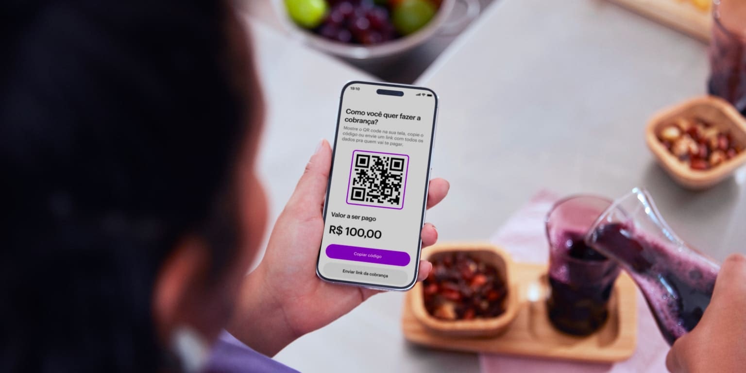 Vista de cima a parte de trás do topo da cabeça de uma mulher que segura na sua mão esquerda seu celular visualizando sua conta no aplicativo do Nubank, enquanto com a outra mão despeja suco de uva de uma jarra transparente em um copo, do lado uma tigela de castanhas e ao fundo uma porção de frutas dentro de uma pia.