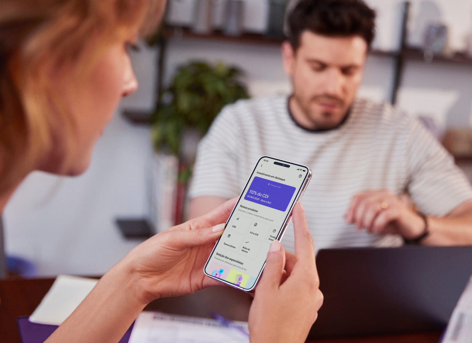 Perfil de uma mulher branca de cabelos curtos segura com as duas mãos um celular e visualiza sua conta no aplicativo do Nubank, ao fundo e desfocado um homem branco olha para um notebook em cima de uma mesa de madeira com um caderno aberto e alguns papéis espalhados.