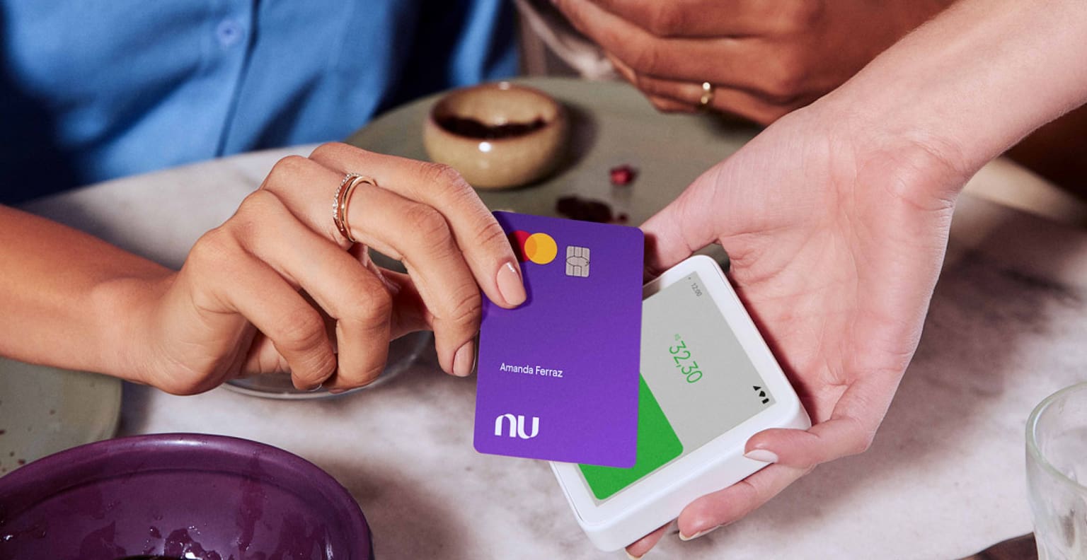 Mulher negra à esquerda usa seu cartão Nubank para pagamento por aproximação enquanto homem à direita passa geleia no pão. Ambos estão sentados à mesa, na qual há uma tigela de açaí pela metade.