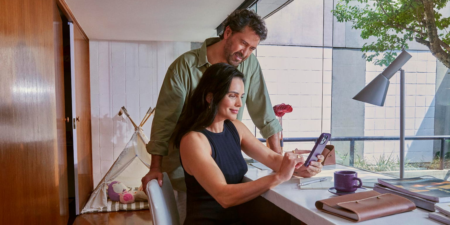 Uma mulher sentada mexendo no celular diante de uma mesa com itens de escritório em casa. Atrás da mulher, um homem, também olhando para o celular que a mulher segura. Ao fundo, uma cabaninha de criança ao lado de um jardim de inverno.