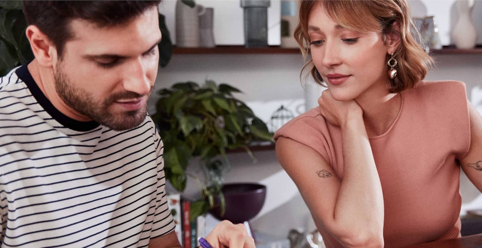 Sentado em uma mesa de madeira um homem branco está lendo alguns papéis enquanto aponta com a caneta para o documento, ao seu lado esquerdo uma mulher branca com a mão esquerda em cima de um caderno observa o que ele está mostrado, a sua frente um notebook, um copo com liquido roxo dentro e uma xícara pequena.
