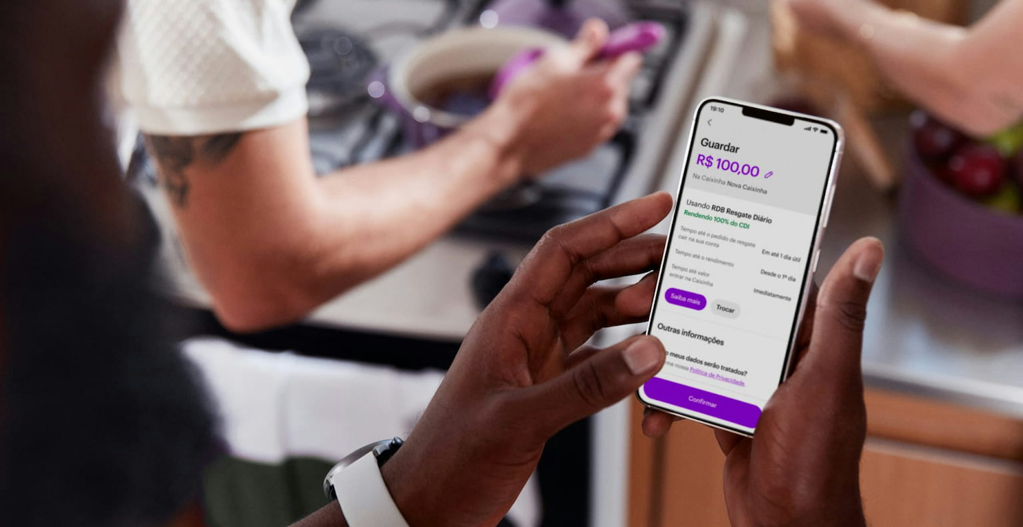 Homem segurando celular com as duas mãos com o aplicativo do Nubank aberto guardando dinheiro nas Caixinhas. Ao fundo há duas pessoas, uma delas cozinhando em uma panela roxa.