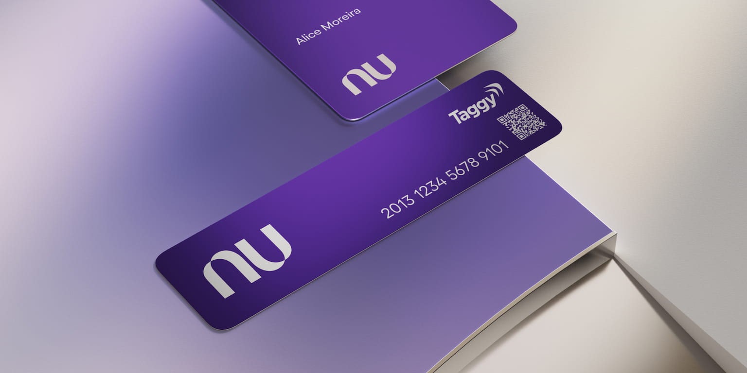 Sobre uma superfície metalizada iluminada por uma luz roxa há um cartão Nubank roxo com o nome Alice Moreira. Abaixo do cartão há uma tag de viagem do Nubank com um QR Code e a marca "Taggy".