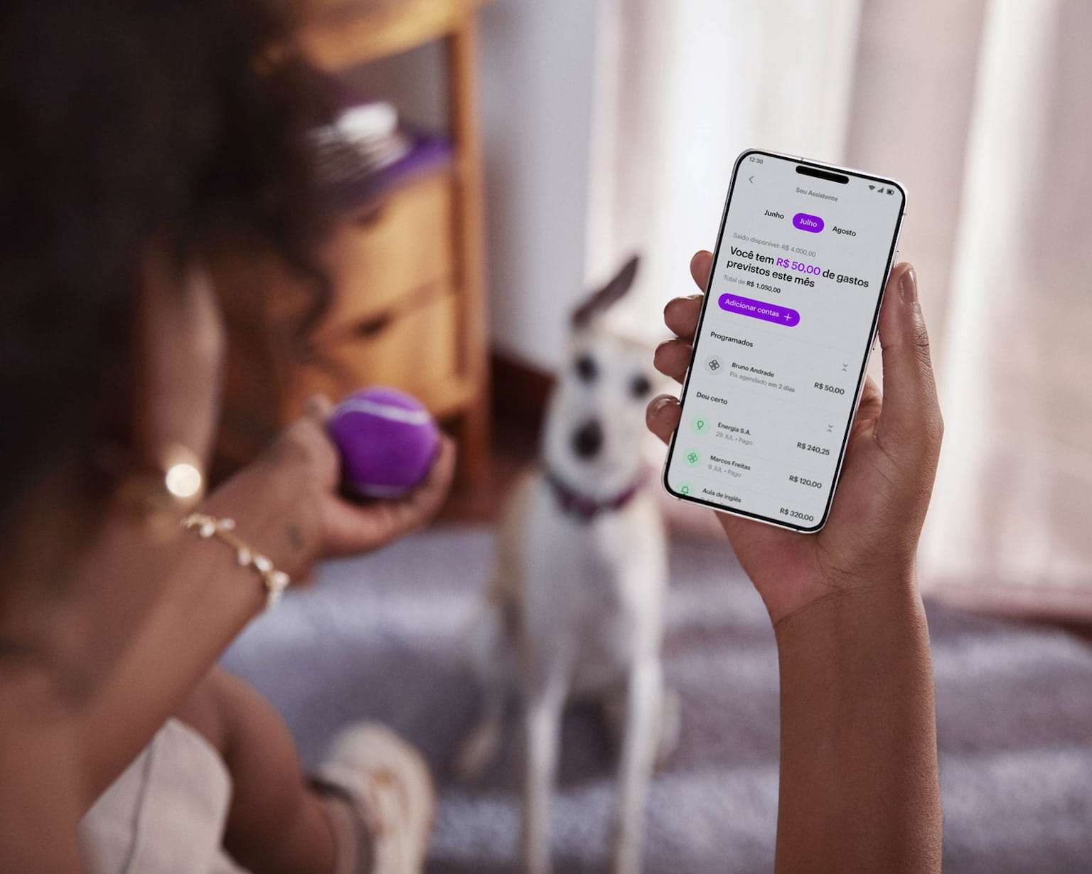 Vista das costas de uma mulher negra sentada de pernas cruzadas, com um celular na mão direita e visualizando sua conta no aplicativo do Nubank, com sua mão esquerda segura uma bolinha de tênis roxa, ao fundo e desfocado um cachorro de pequeno porte sentado no chão olhando para a bolinha.