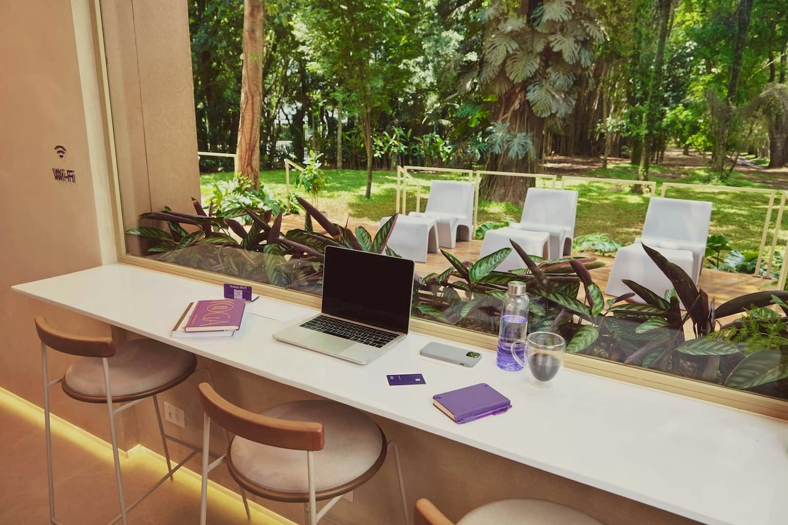 Imagem do lado de dentro da Casa Nubank Ultravioleta – a estação de trabalho, com um balcão branco e amadeirado, com bancos de madeira clara e de frente para uma janela com cadeiras de descanso.