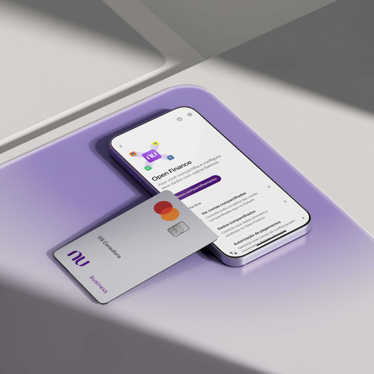 Sobre uma superfície de vidro quadrada roxa há um smartphone na seção de Open Finance no App Nubank. Apoiado sobre ele, há um Cartão Business branco ou Cartão PJ Nubank com nome GS Consultoria.