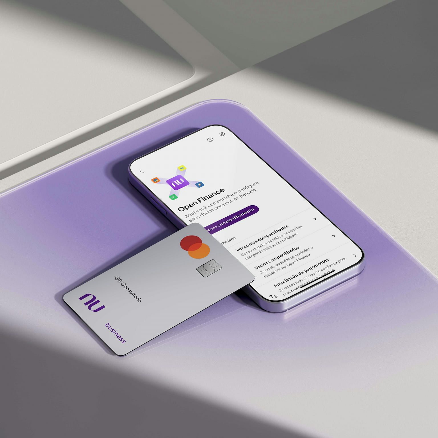 Sobre uma superfície de vidro quadrada roxa há um smartphone na seção de Open Finance no App Nubank. Apoiado sobre ele, há um Cartão Business branco ou Cartão PJ Nubank com nome GS Consultoria.