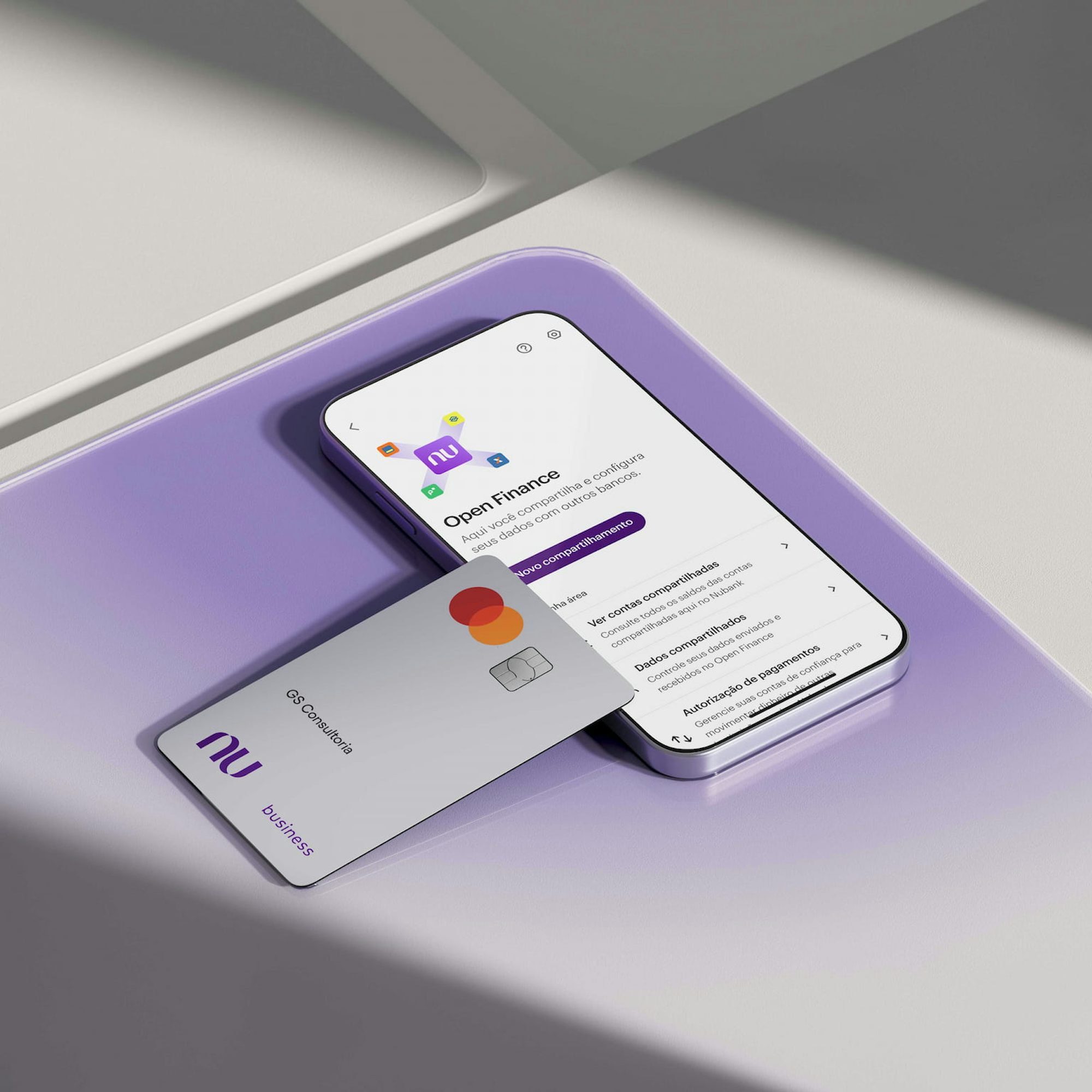 Sobre uma superfície de vidro quadrada roxa há um smartphone na seção de Open Finance no App Nubank. Apoiado sobre ele, há um Cartão Business branco ou Cartão PJ Nubank com nome GS Consultoria.