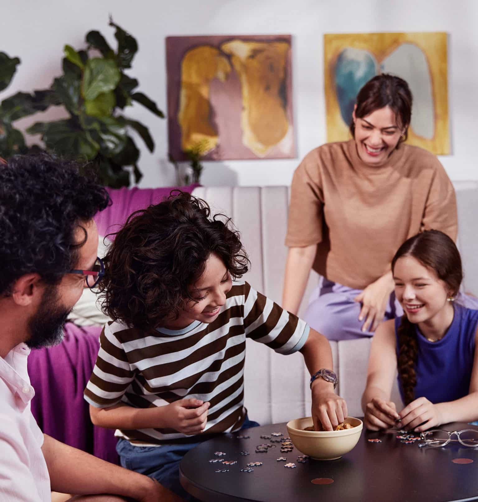 Quatro pessoas em uma sala, homem sentado no chão olha para duas crianças sorrindo e sentadas no chão em volta de uma mesa de centro enquanto brincam com peças de um quebra-cabeça, ao fundo uma mulher sentada no sofá observa as crianças enquanto sorri.