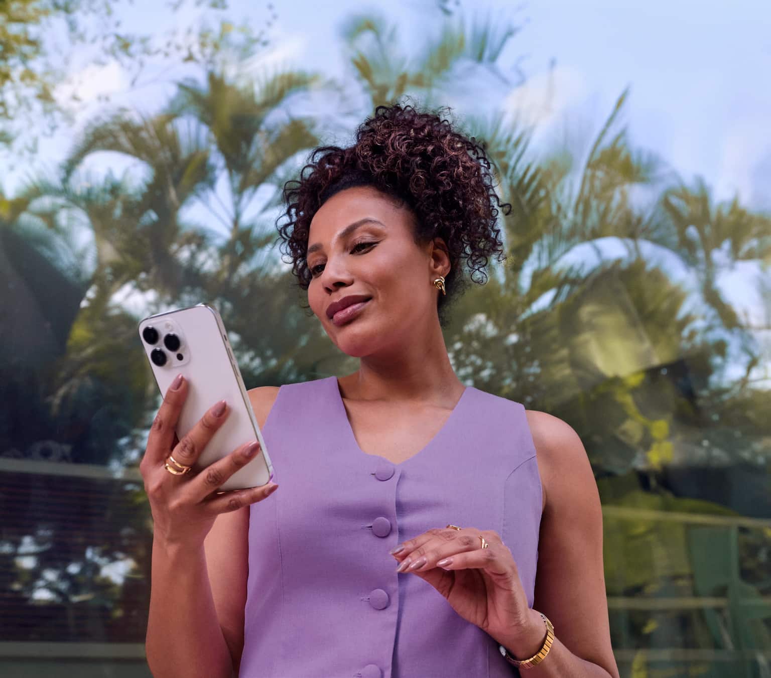 Mulher negra, com cabelos cacheados, presos, está de pé ao ar livre, próxima a palmeiras. Ela veste um colete roxo, com três botões, segura um celular e está olhando para ele.