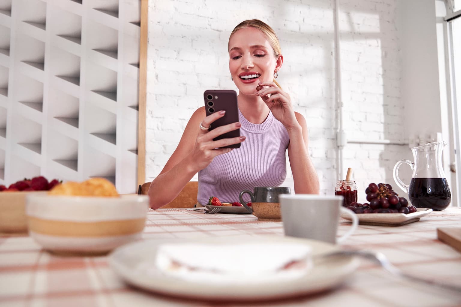 Mulher branca com o cabelo preso, sentada atrás de uma mesa com xícaras e tigelas com frutas e uma jarra de suco, olha para o celular que está em uma de suas mãos sorrindo. Com a outra mão, leva uma uva a sua boca. Ao fundo uma parede de tijolos brancos com um quadro abstrato iluminados pela luz do dia de parte e uma janela.