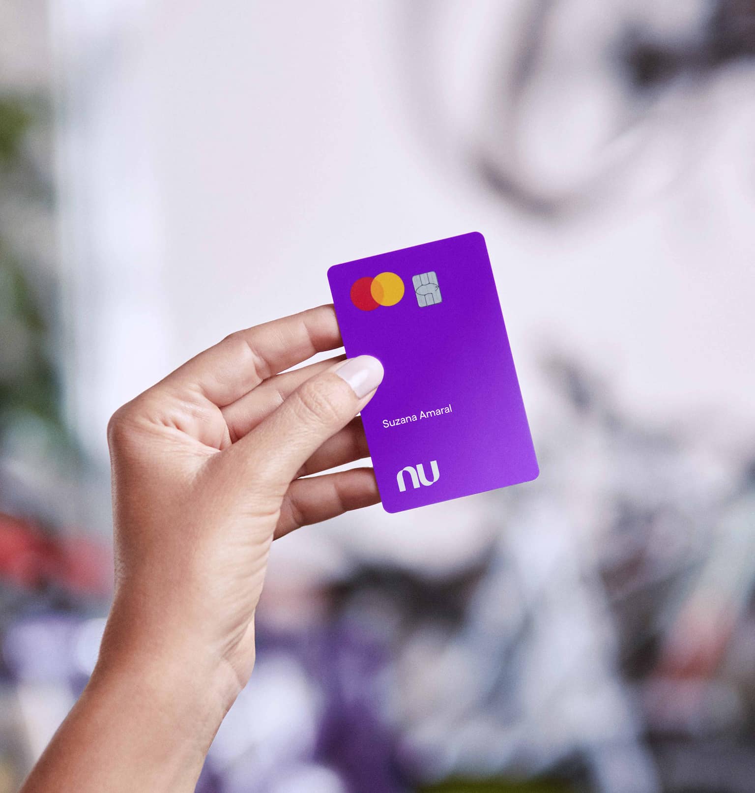 Uma mão de mulher branca segura o Cartão Nubank roxo. Essa mão está centralizada na imagem e em primeiro plano.