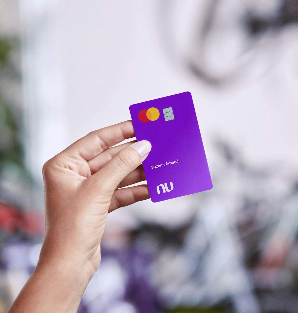 Uma mão de mulher branca segura o Cartão Nubank roxo. Essa mão está centralizada na imagem e em primeiro plano.