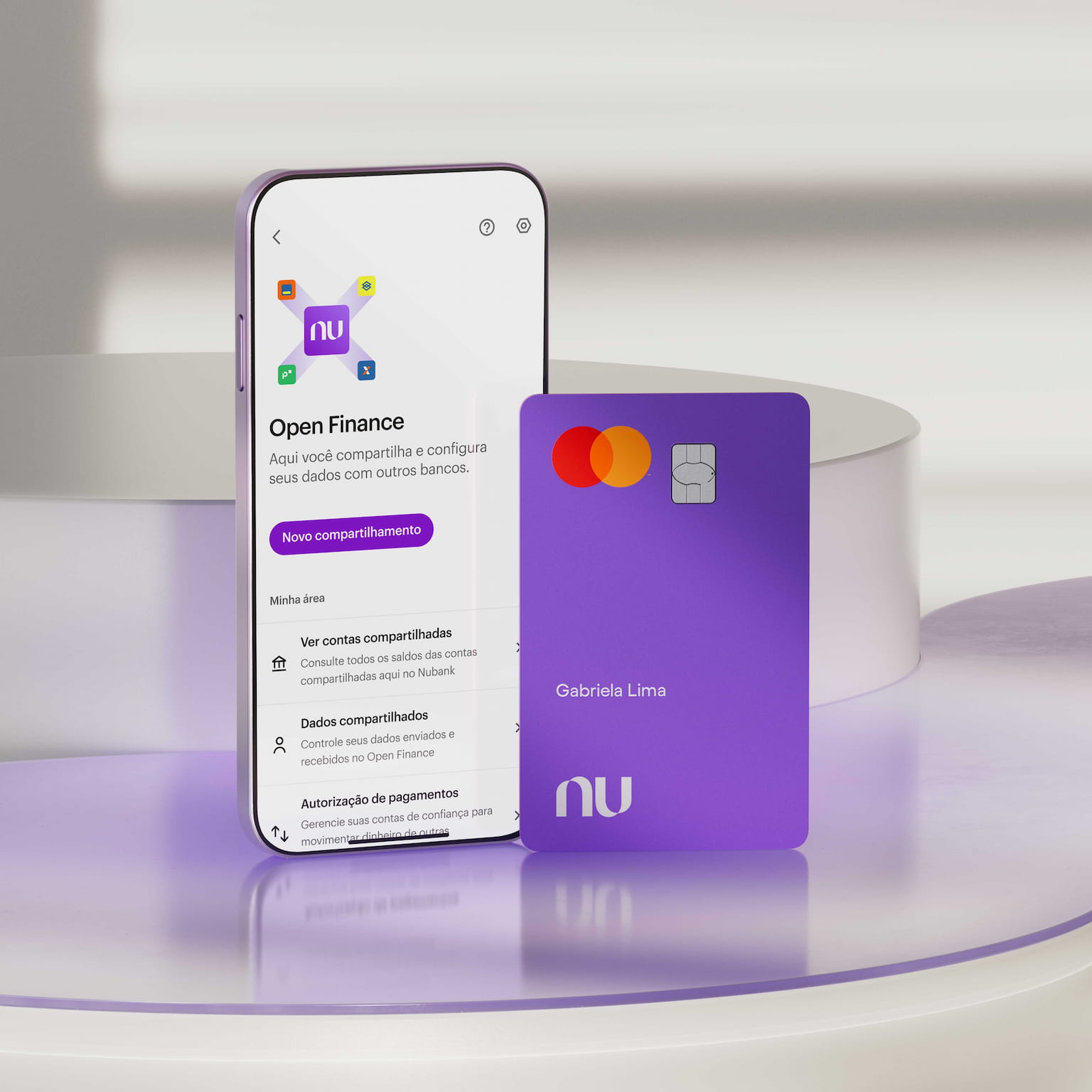 Sobre uma superfície de vidro roxa e apoiados em um bloco redondo branco, há um smartphone na seção de Open Finance em sua tela e um cartão roxo do Nubank com o nome Gabriela Lima.