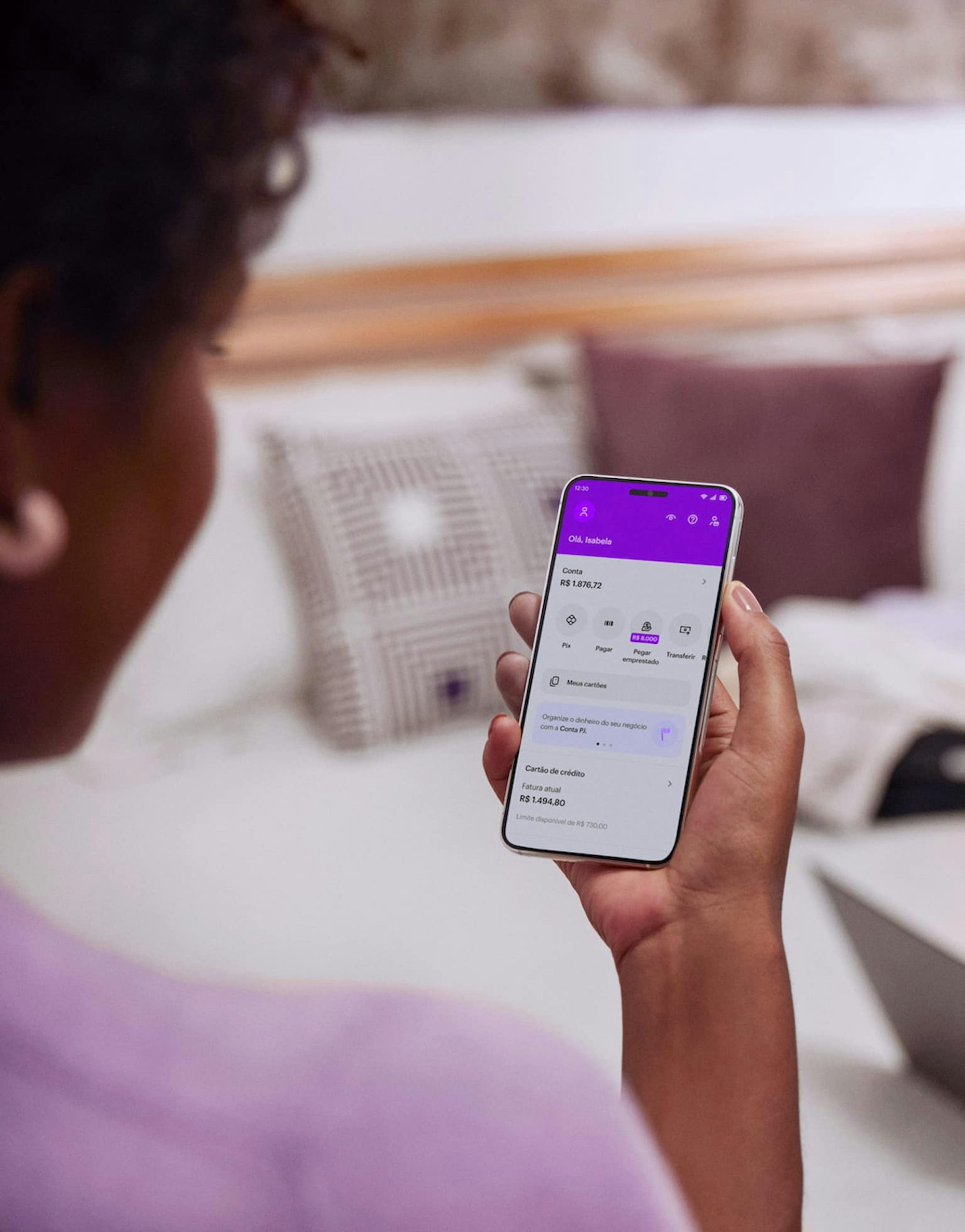 Vista das costas de uma mulher negra sentada com um celular na mão direita e visualizando sua conta no aplicativo do Nubank.