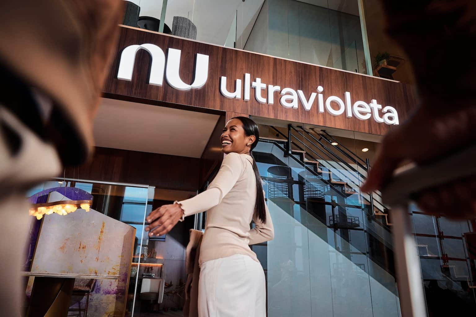 Mulher negra sorrindo, usando uma roupa em tons claros, em frente ao Nubank Ultravioleta Lounge, no Aeroporto de Guarulhos.