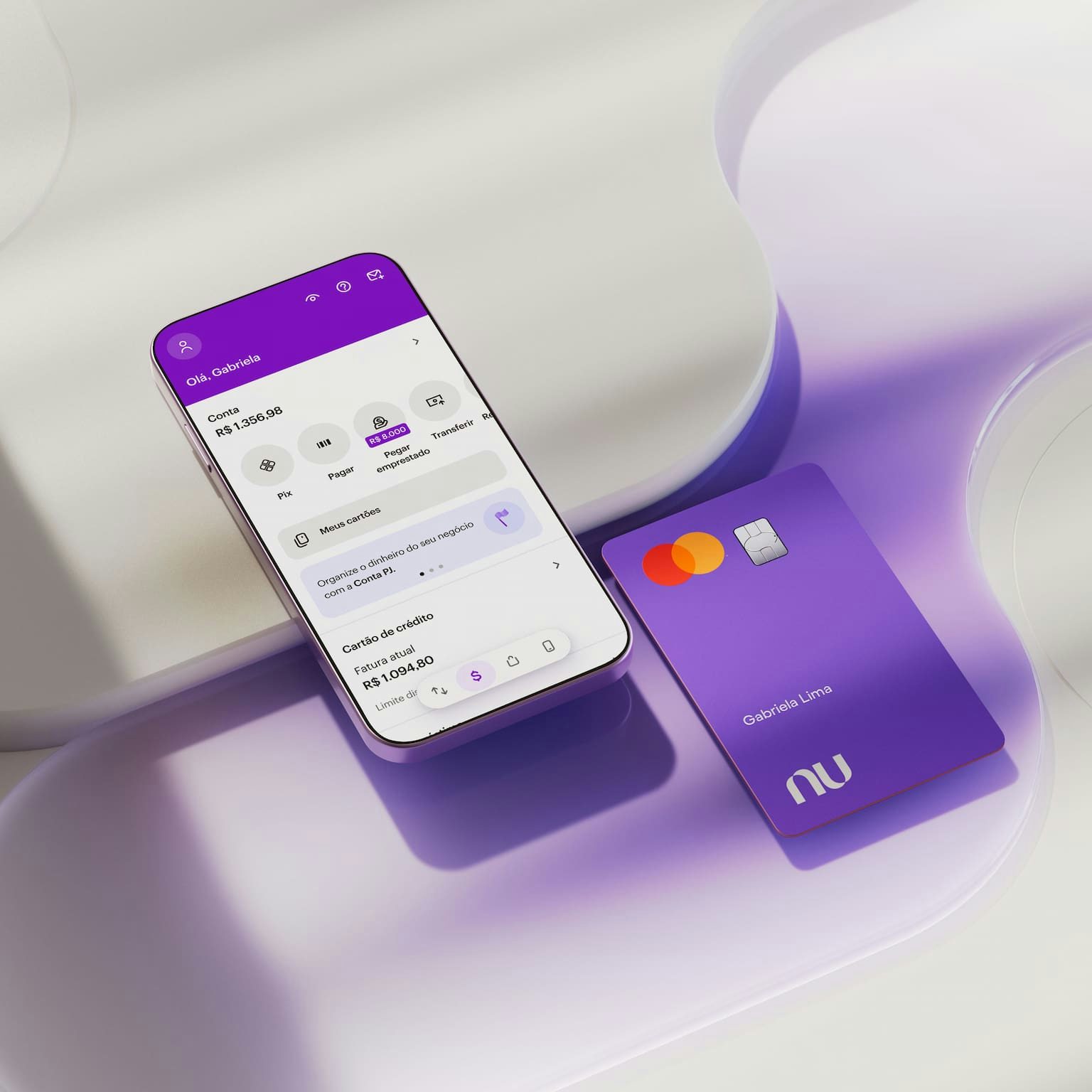 Celular com aplicativo Nubank aberto na área inicial com o saldo da conta na parte superior da tela. Na parte inferior do aplicativo na tela do celular, informações sobre Cartão de crédito. O celular e o cartão estão deitados apoiados em uma forma geométrica branca.