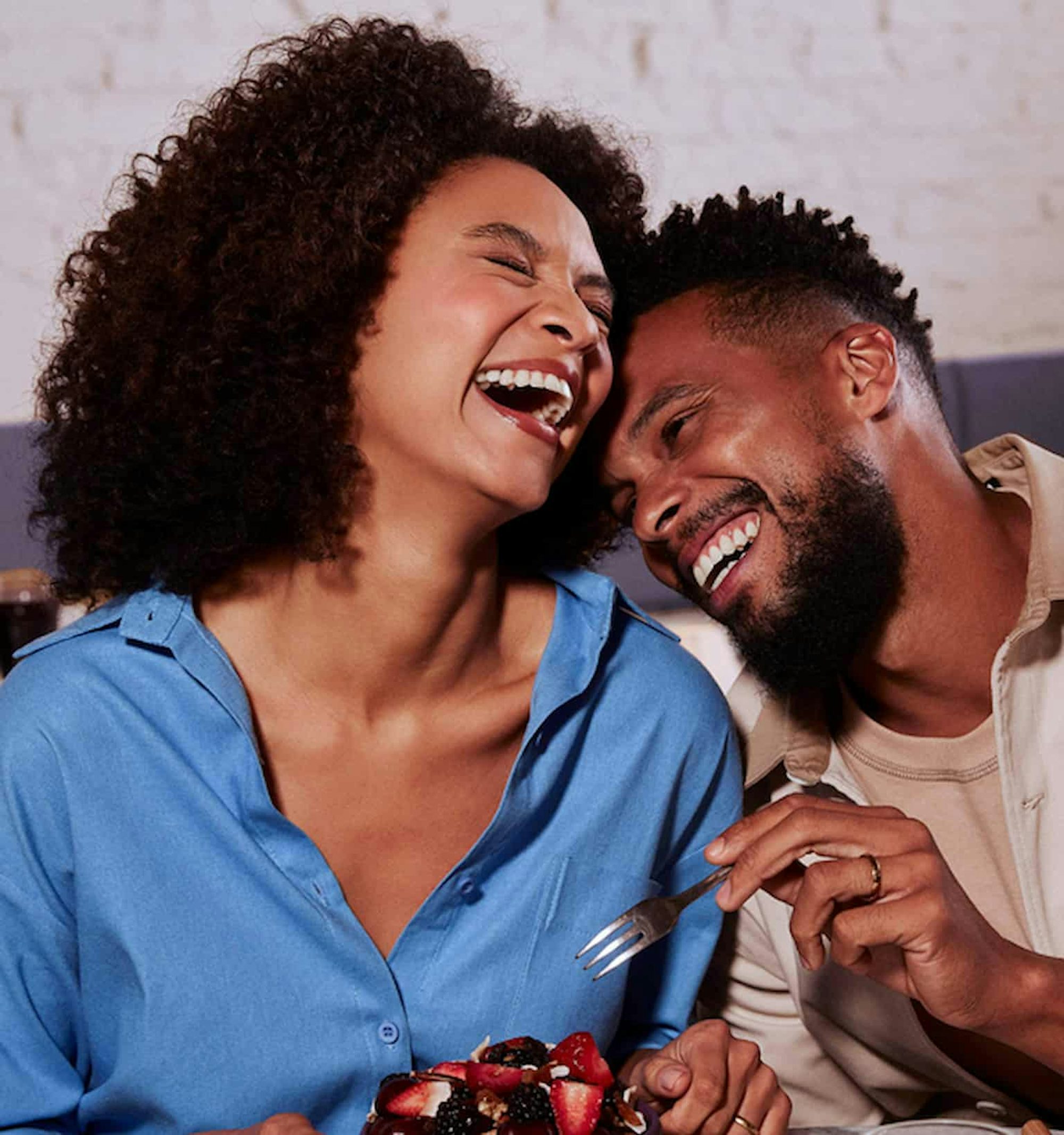 Uma mulher negra à esquerda ri segurando uma tigela com frutas. À sua direita um homem negro ri encostado nela com um garfo em direção à sua tiigela. Ambos estão sentados à uma mesa de café da manhã.