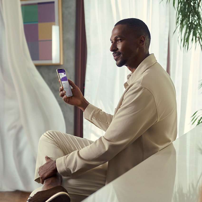 Homem negro usando roupas claras. Ele está em uma sala com cortinas brancas e está segurando um celular com uma das mãos.