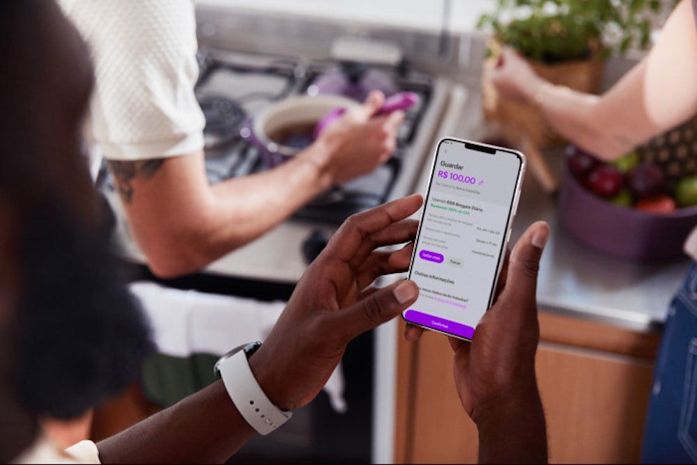 Homem segurando celular com as duas mãos com o aplicativo do Nubank aberto guardando dinheiro nas Caixinhas. Ao fundo há duas pessoas, uma delas cozinhando em uma panela roxa e a outra pessoa manuseia frutas.
