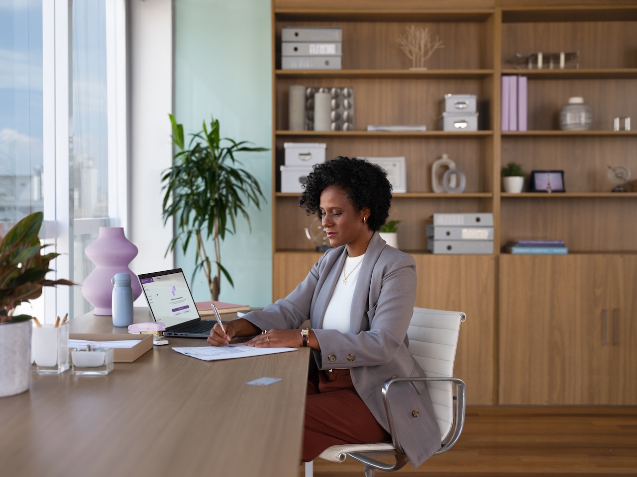 Mulher empreendedora trabalha em um escritório moderno e bem iluminado. Ela está sentada à mesa, escrevendo em um papel, com um notebook aberto à sua frente, exibindo a interface do Nu Empresas. Ao fundo, há uma estante de madeira com objetos decorativos e materiais organizados, transmitindo um ambiente profissional e acolhedor.