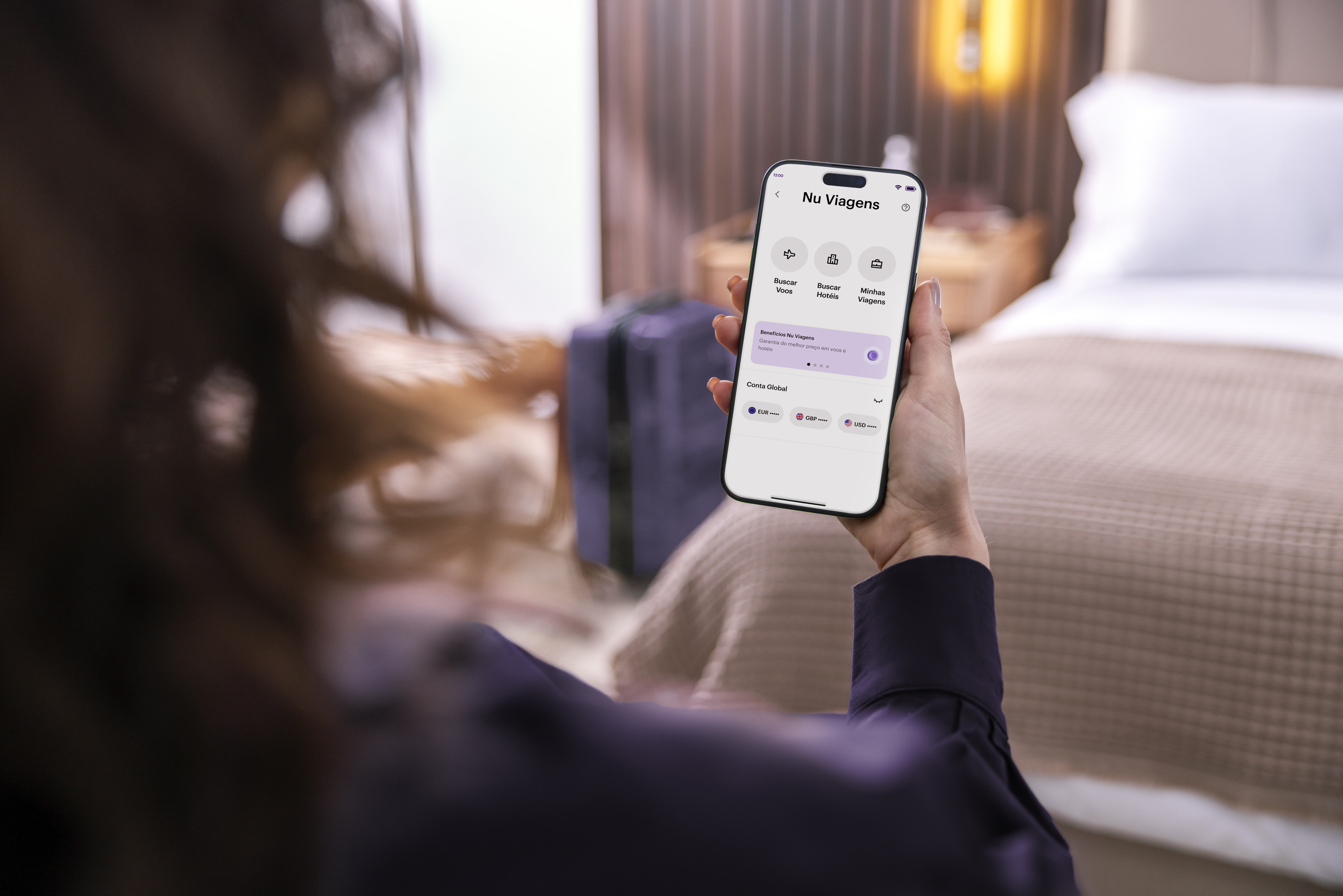 Mulher branca sentada no quarto de um hotel olhando a tela do seu celular com a tela do Nu Viagens
