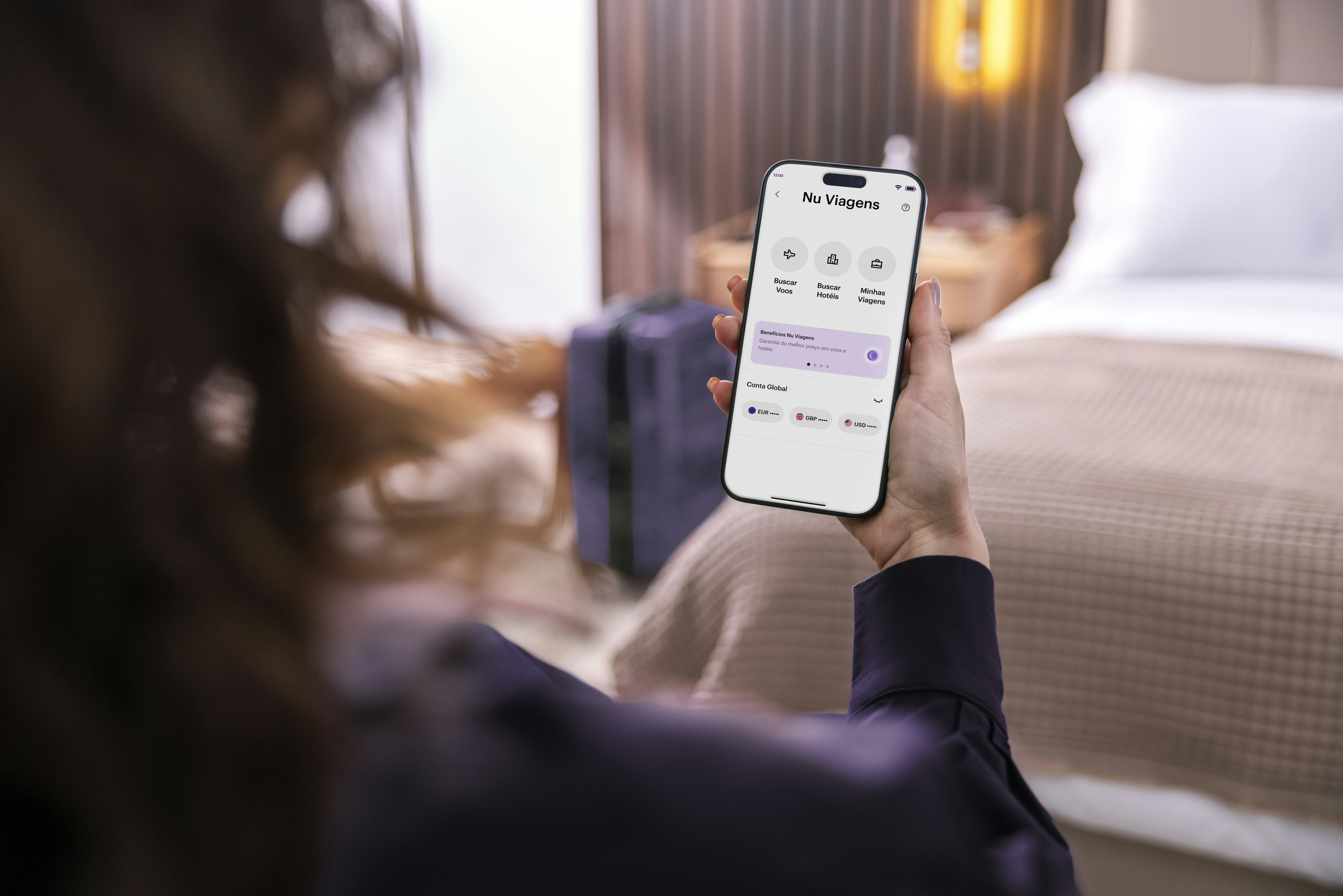 Mulher branca sentada no quarto de um hotel olhando a tela do seu celular com a tela do Nu Viagens
