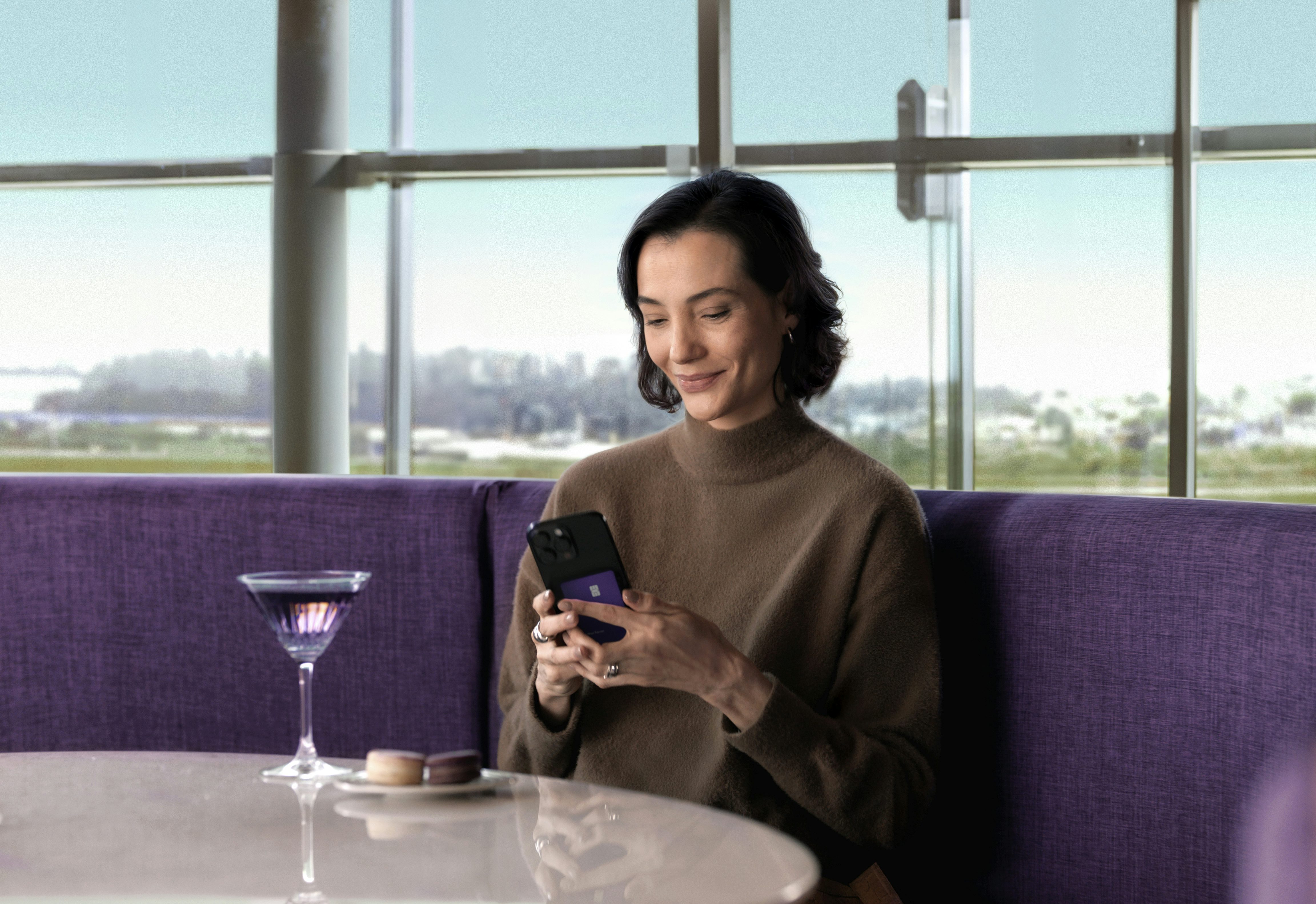 Mulher branca de carlos curtos sentada em um sofá roxo em frente a uma mesa com um drink também roxo olhando sorrindo para o seu celular