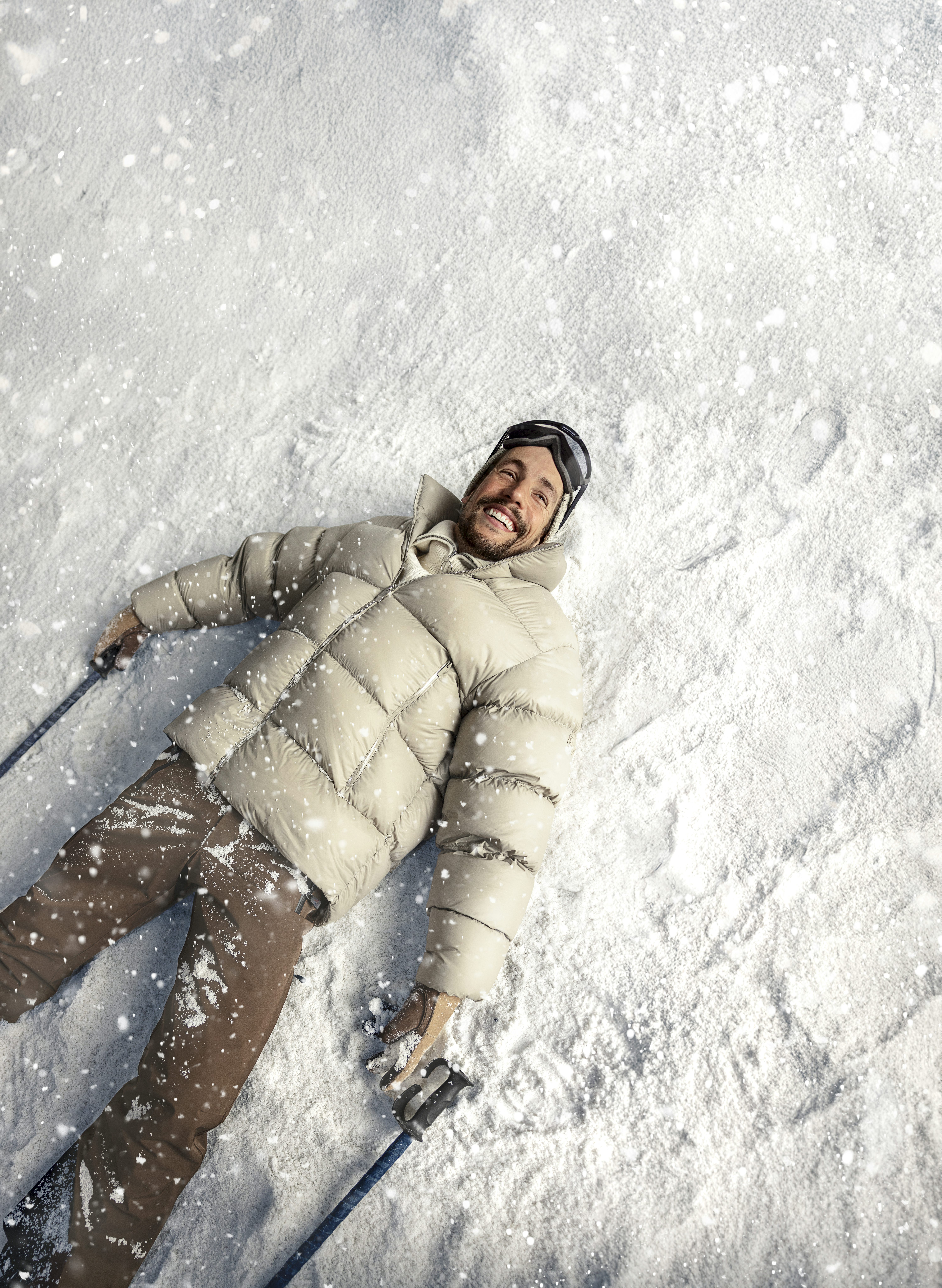 Homem branco deitado na neve sorrindo com roupas de ski.