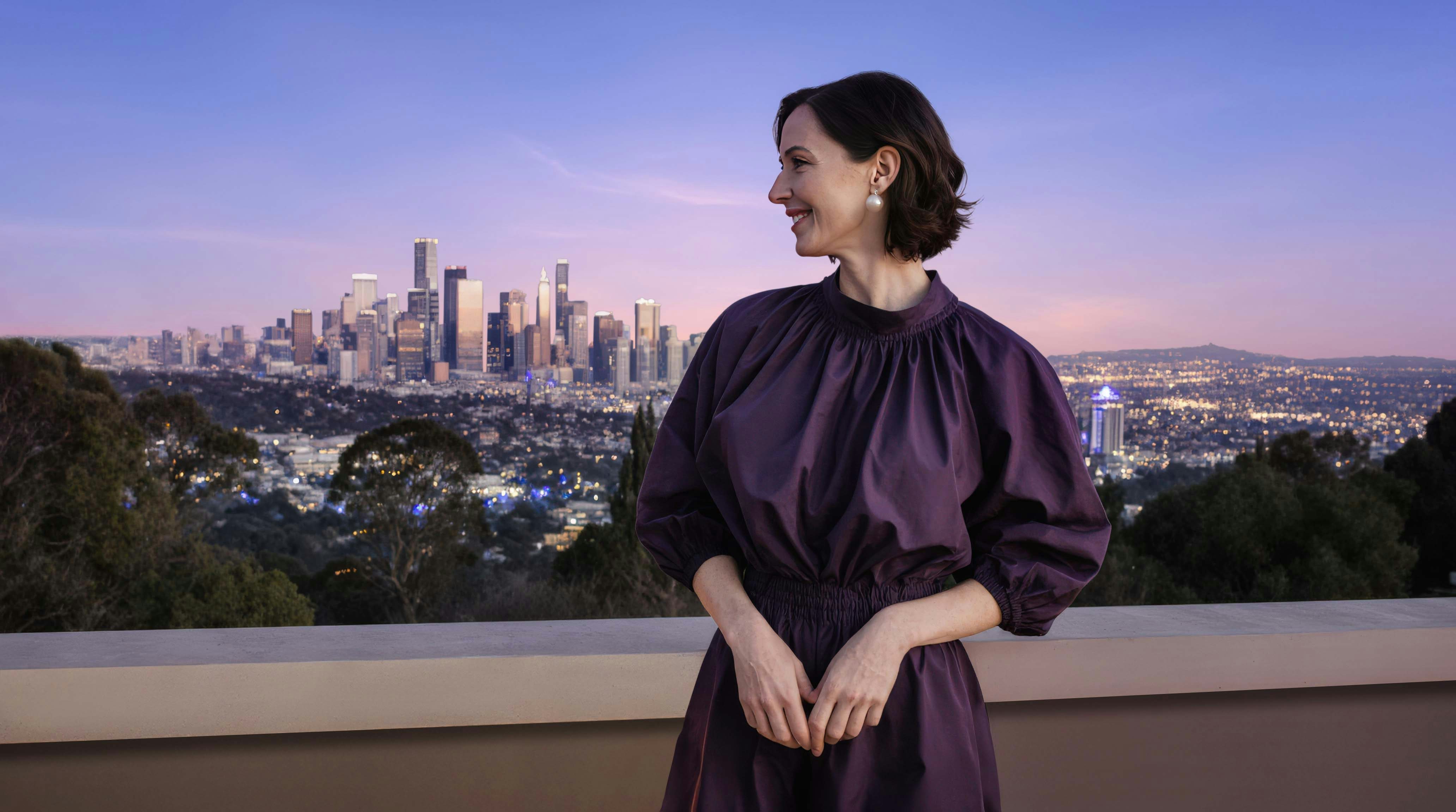 Mulher branca tranquila e sorrindo olhando ao horizonte o que parece ser uma cidade como Los Angeles
