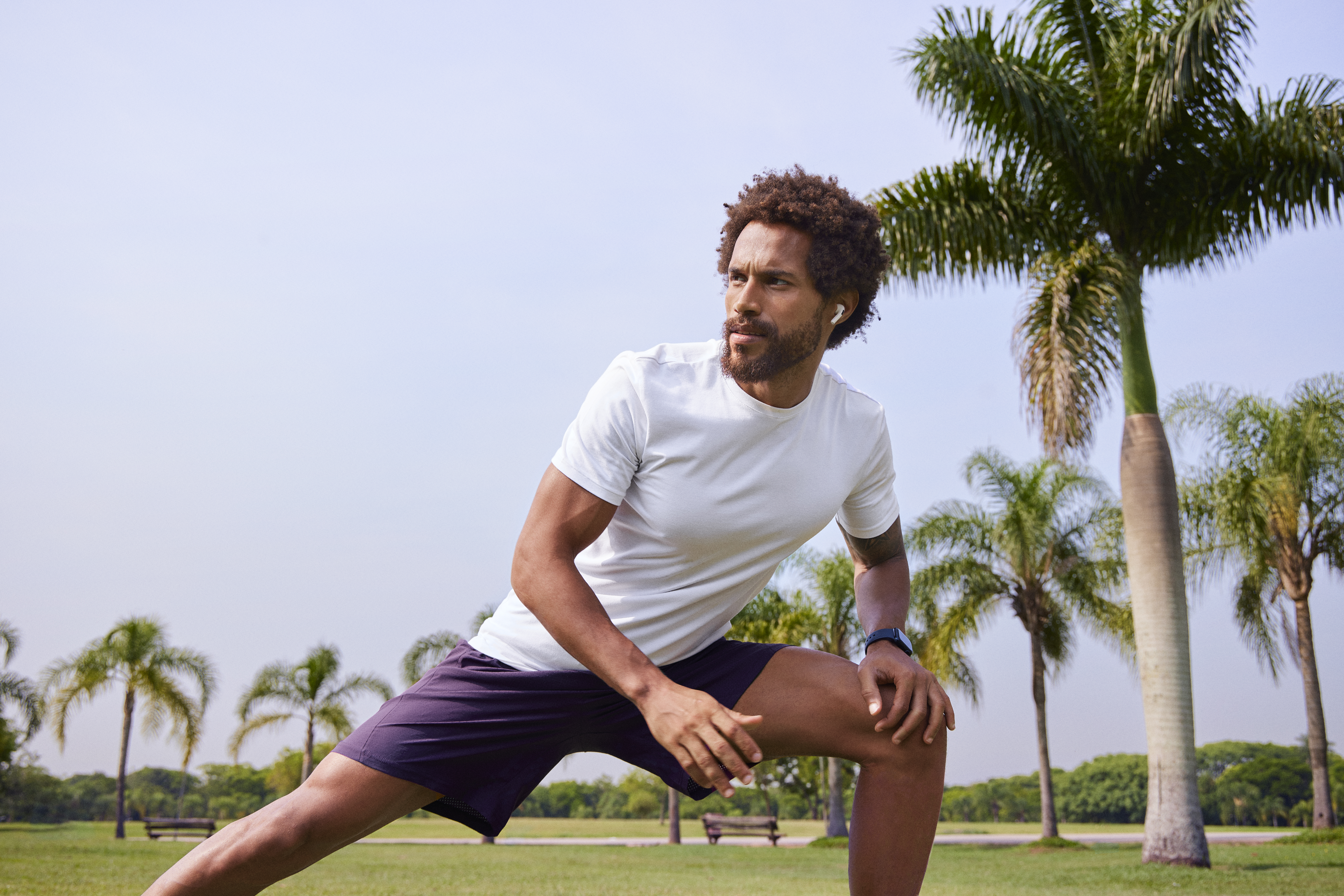 Homem negro se alongando durante exercício em um parque, com palmeiras ao fundo, vestindo camiseta branca, shorts escuros e fones de ouvido.