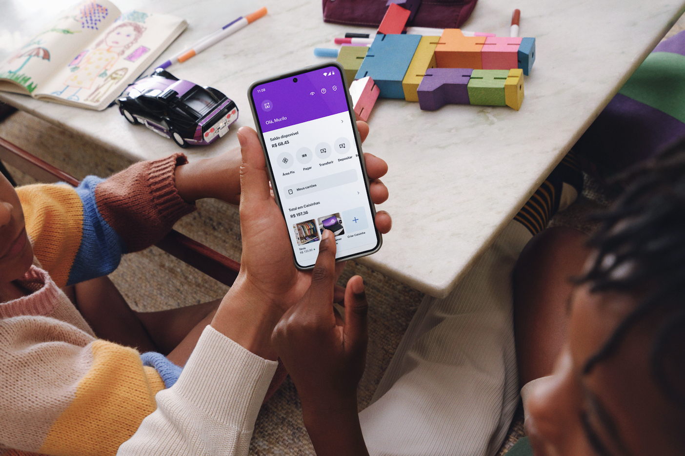 Duas crianças sentadas junto com um adulto mexendo no celular com o aplicativo do Nubank e ao fundo uma mesa com brinquedos sobre ela.