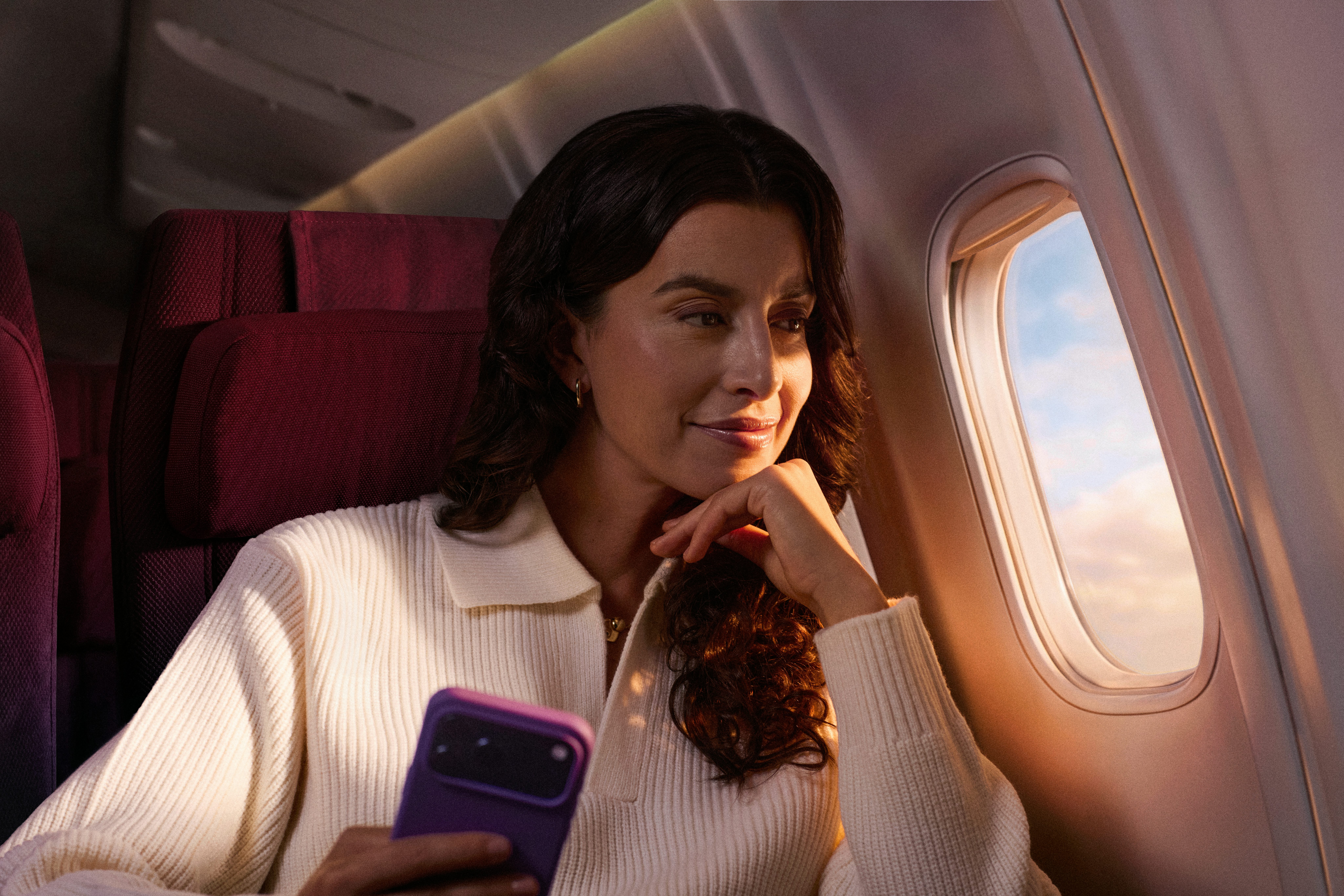 Mulher com cabelos soltos segurando um celular. Ela está perto da janela do avião e usa uma blusa de tricô branca.
