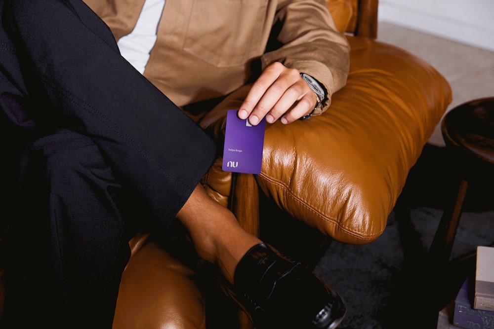 Detalhe em ângulo mais fechado de um homem sentado em uma poltrona de couro caramelo com as pernas cruzadas. Ele segura o cartão Nubank Ultravioleta sobre o braço da poltrona.