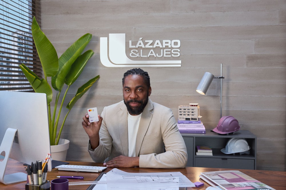 Lázaro Ramos sentando no escritório do seu novo empreendimento Lázaro & Lajes segura o cartão Nu Empresas com uma das mãos