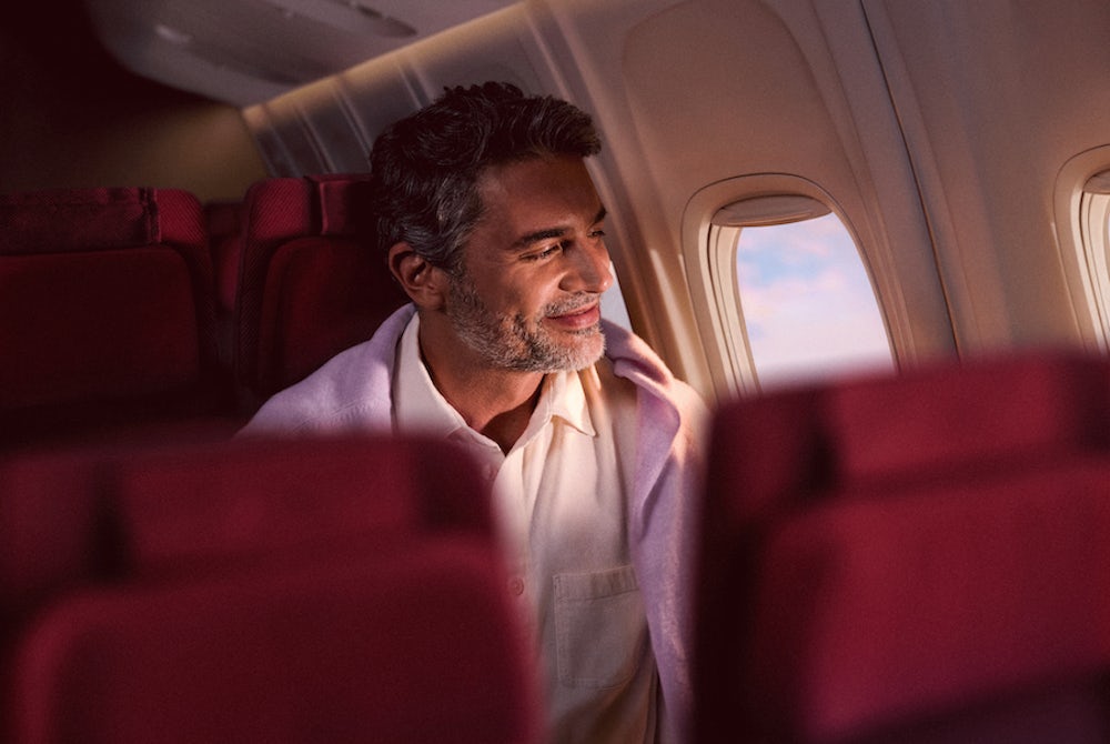 Um homem grisalho sorri enquanto olha pela janela de um avião durante o voo. Ele está coberto por uma manta lilás clara, acomodado em uma poltrona de tecido vinho.