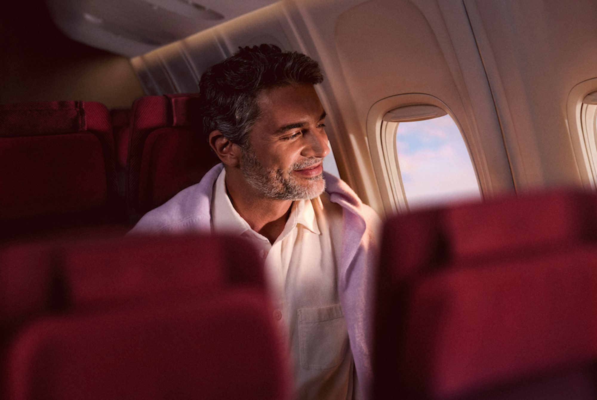 Um homem grisalho sorri enquanto olha pela janela de um avião durante o voo. Ele está coberto por uma manta lilás clara, acomodado em uma poltrona de tecido vinho.