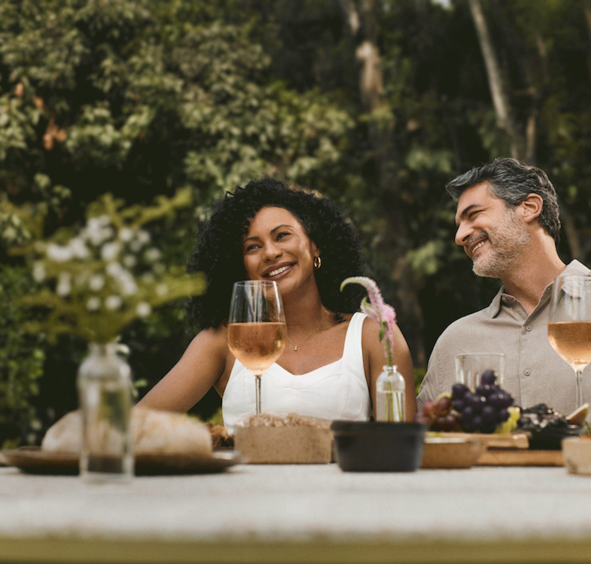 Uma mulher negra e um homem branco rindo, sentados à mesa, com uvas e taças de vinho rosé.